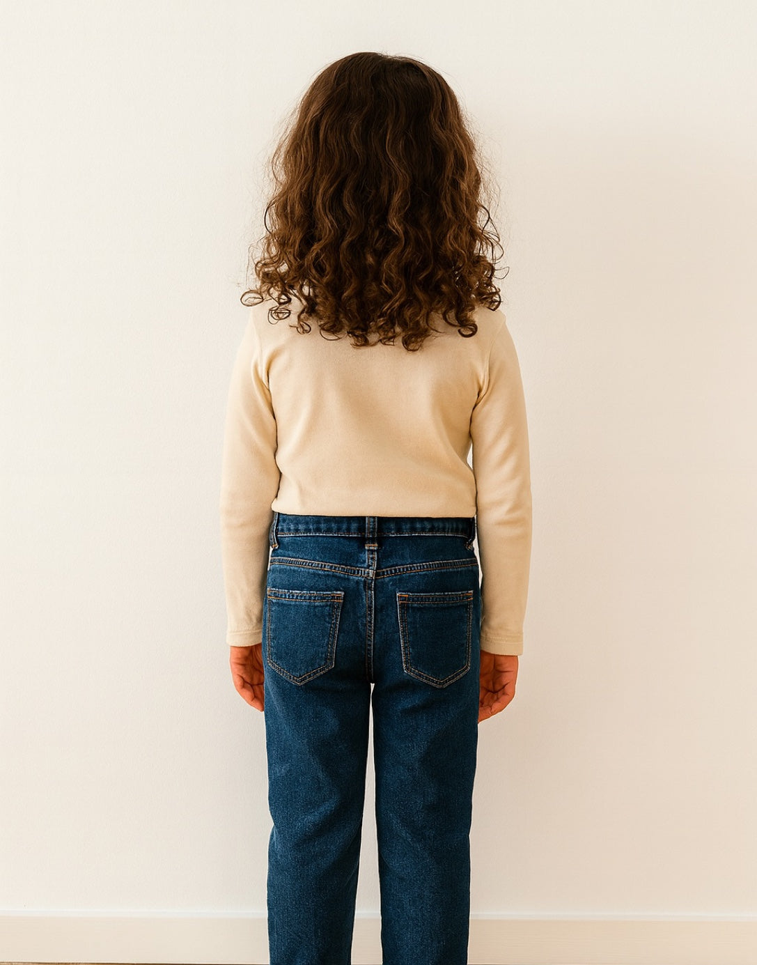 Back view of young girl wearing a beige Long Sleeve Scoop Neck Top and dark jeans against a white wall