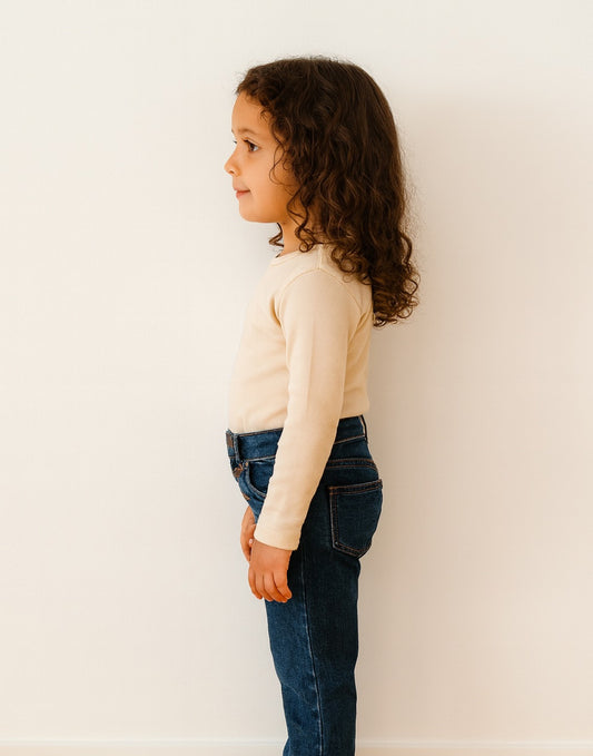 Side view of young girl wearing a beige Long Sleeve Scoop Neck Shirt and dark jeans against a white wall