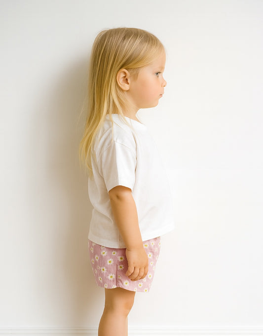 Side view of young girl wearing a white top and pink floral shorts against a white background.