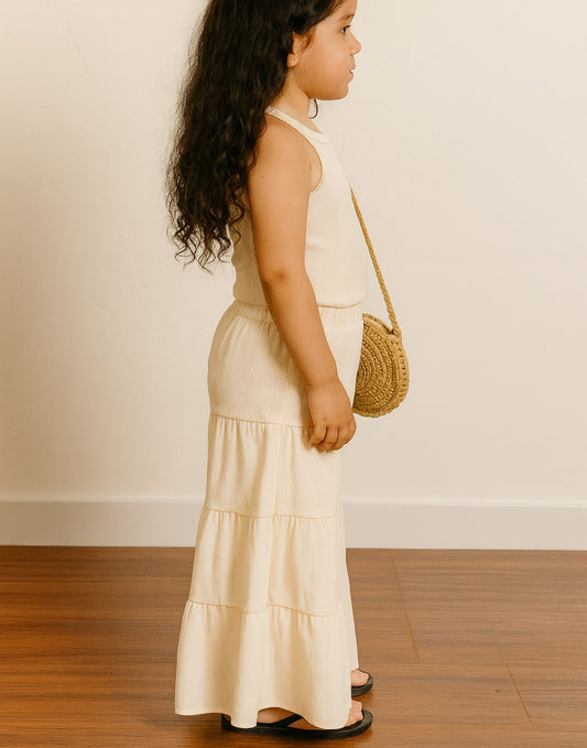 Side view of young girl in a beige Tiered Summer Pants holding a round straw bag against a plain background.