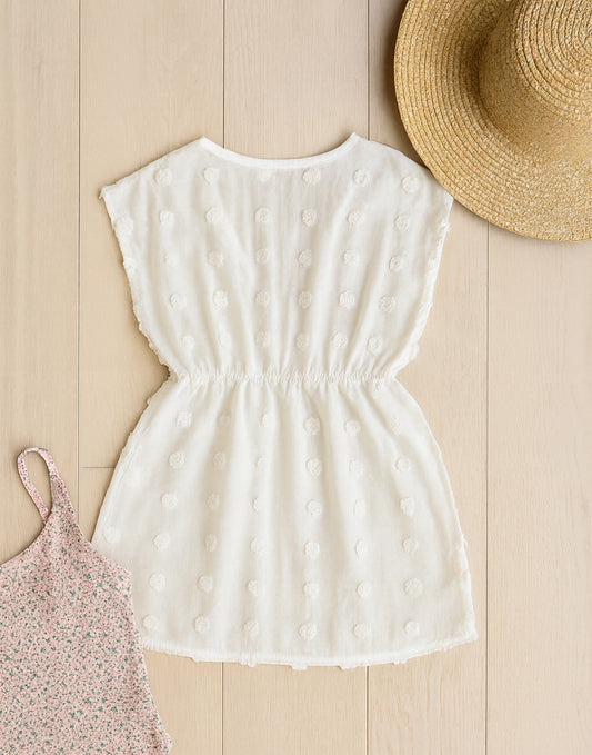 Back view of white polka dot Girls Swimsuit Tunic Coverup on a wooden surface with a straw hat and pink floral dress.