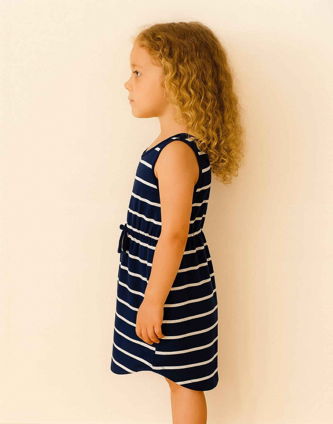 Side view of young girl wearing a navy and white striped Tie Front Curved Hem Knit Dress against a plain background.