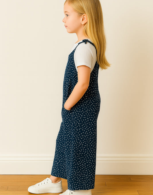 Side view of young girl wearing a navy blue polka dot Tie Shoulder Jumpsuit with white sneakers on a plain background
