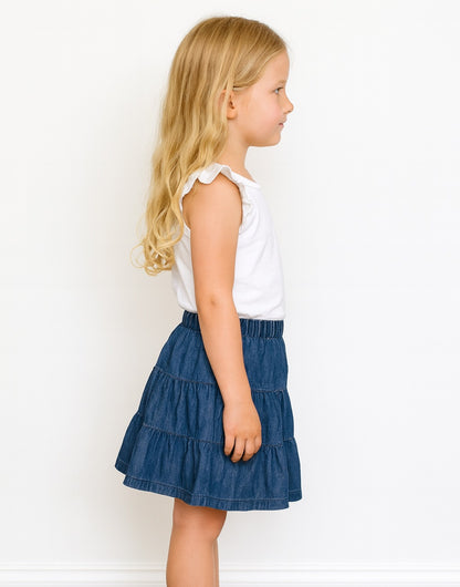 Side view of young girl wearing a white top and blue denim Tiered Mini Skirt on a white background.