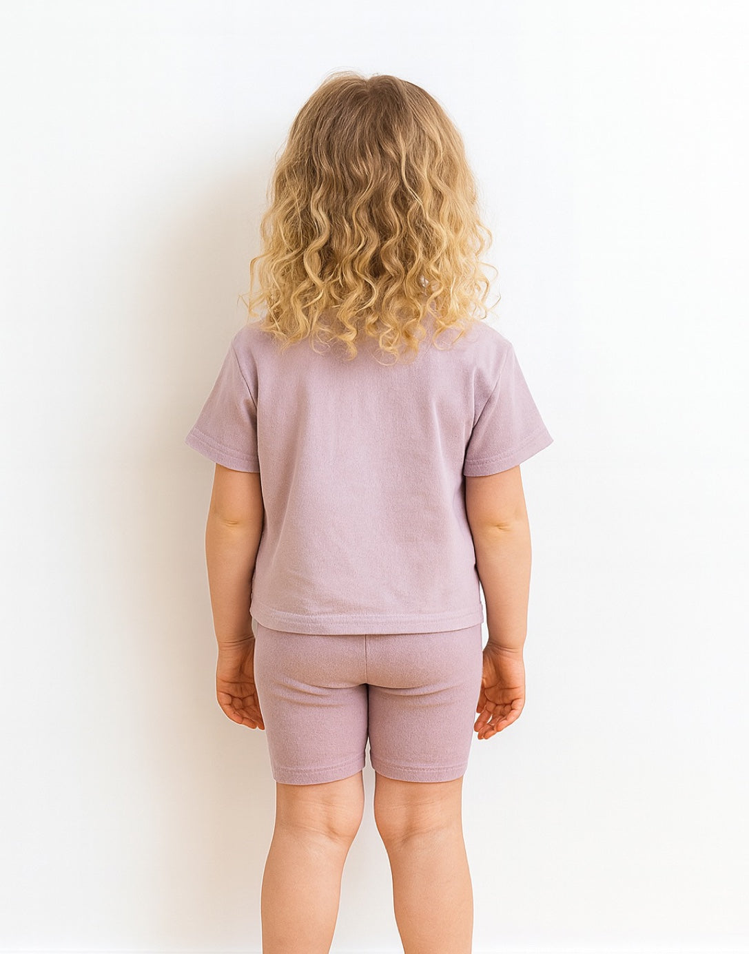 Back view of young child wearing a light purple t-shirt and shorts set on a white background.