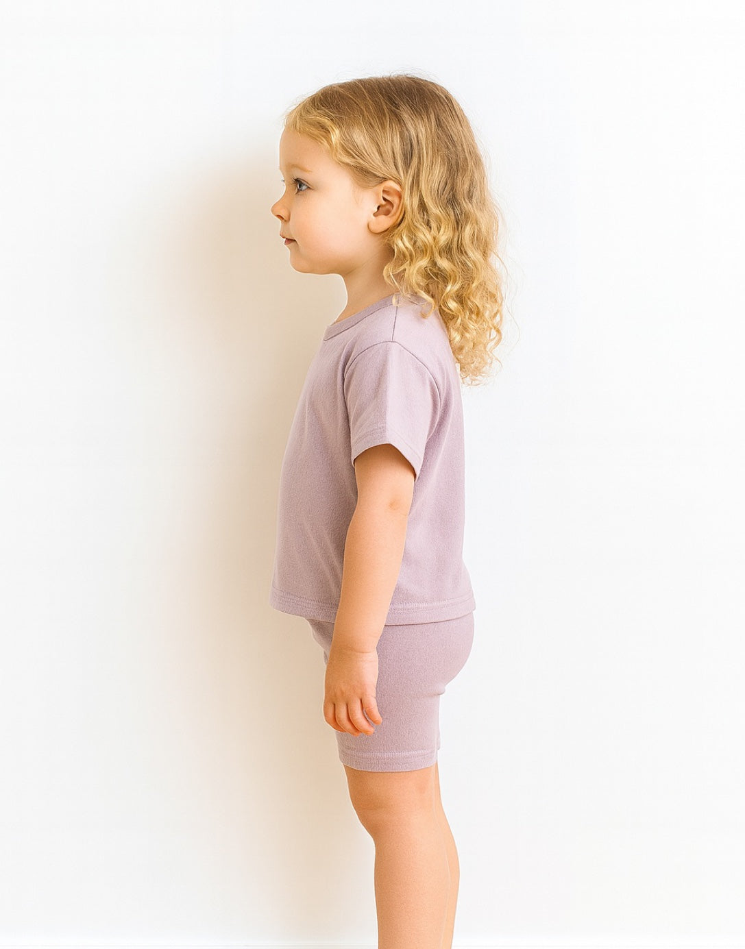 Side view of young child wearing a light purple t-shirt and shorts set on a white background