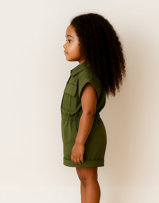 Side view of young girl in a green Utility Romper standing in front of plain white background