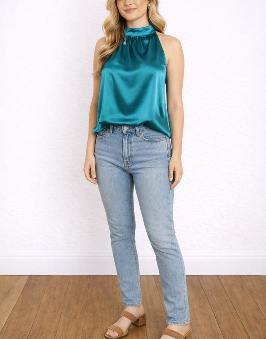 Woman wearing a teal Halter Neck Top and blue jeans against a white wall.