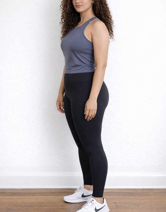 Side view of woman wearing a gray Halter Tank Top and black leggings against a white wall.