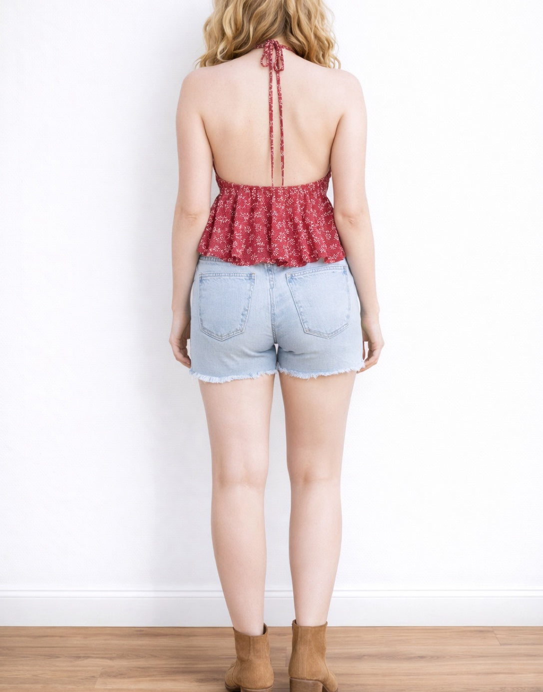 Back view of woman wearing a red floral Halter Tie Back Crop Top and denim shorts against a white background.