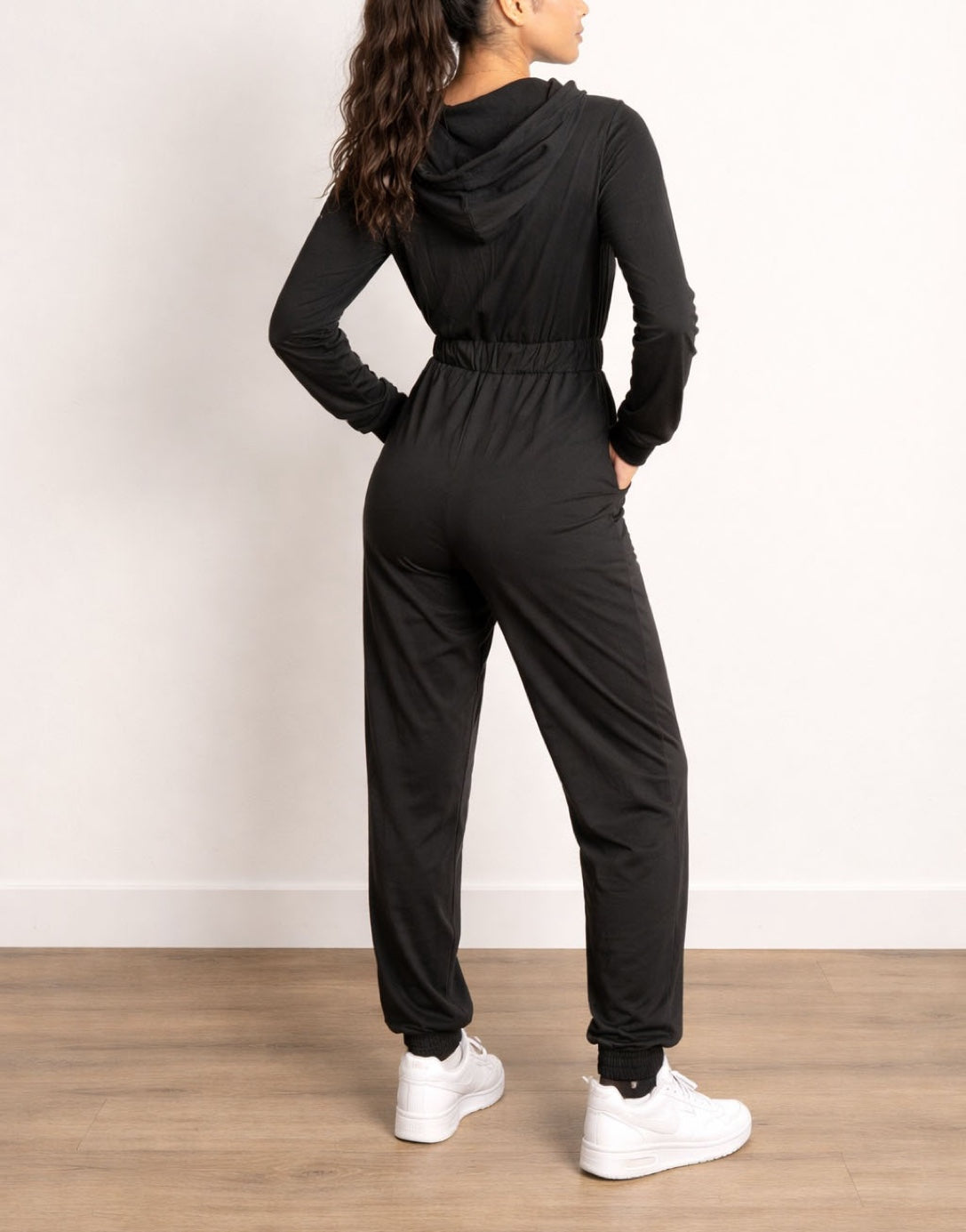 Back view of woman wearing a black Hooded Jumpsuit with white sneakers on a wooden floor.