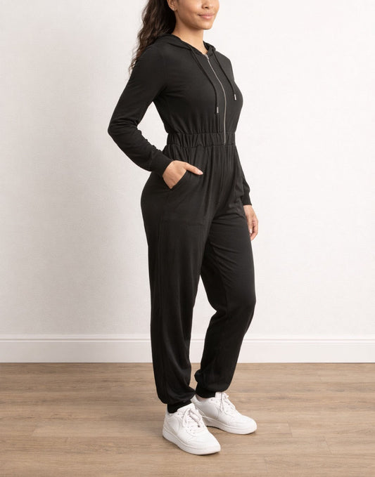 Side view of woman wearing a black Zip Up Hooded Jumpsuit with white sneakers on a wooden floor.