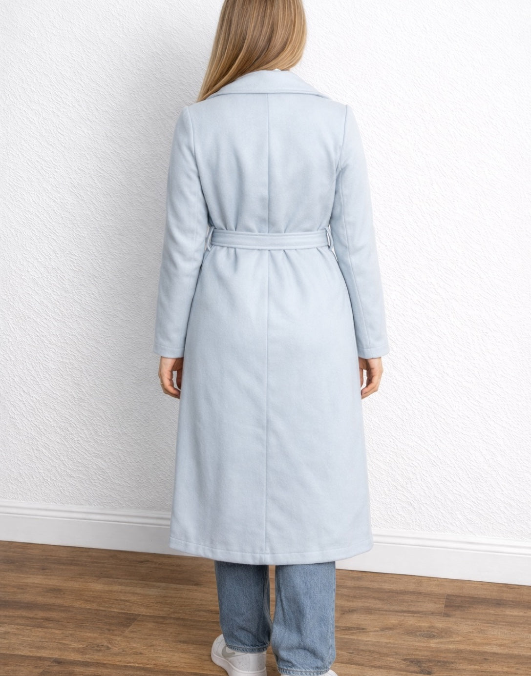Back view of woman wearing a light blue Long Belted Coat standing against a white wall.