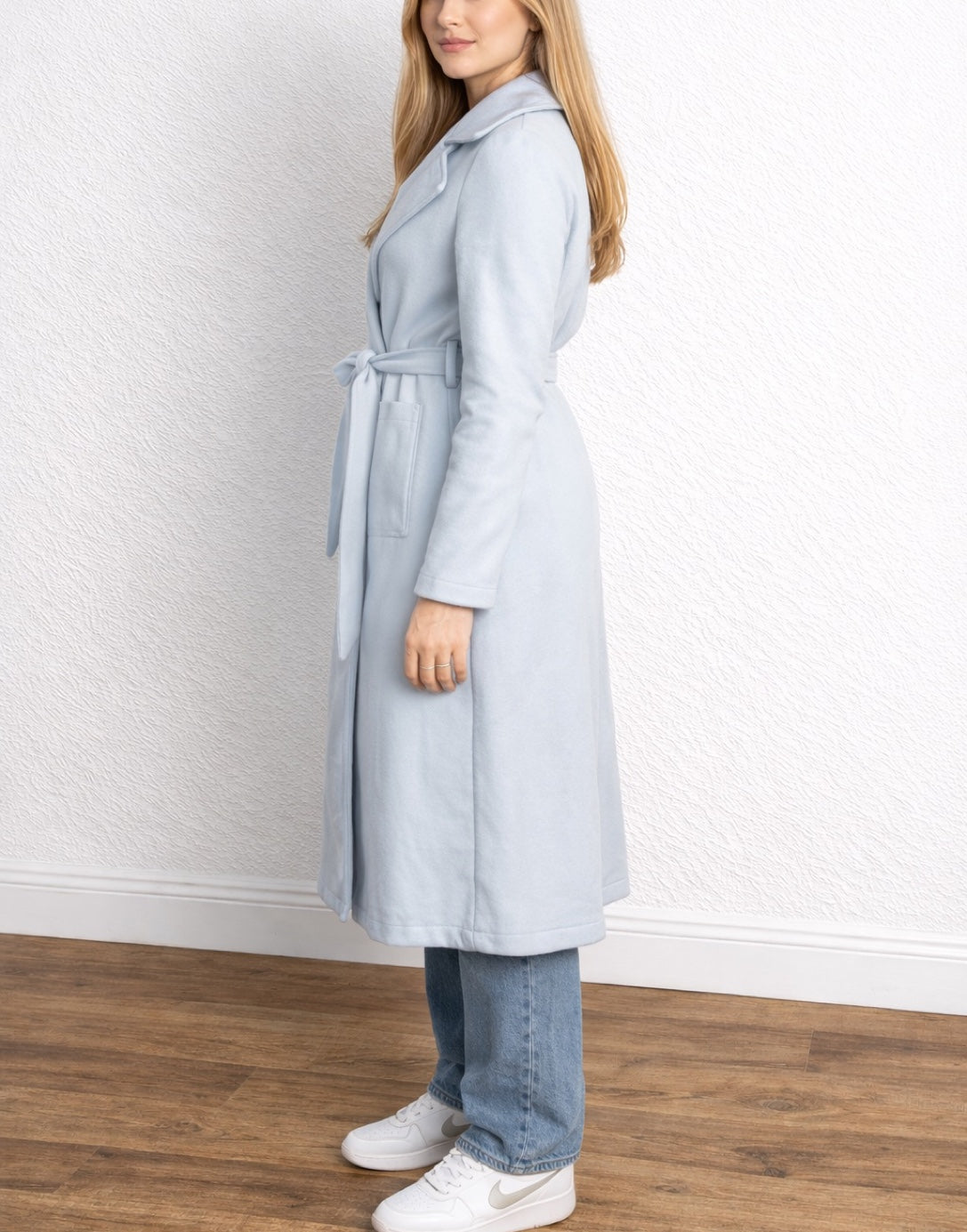 Side view of woman wearing a light blue Long Belted Coat standing against a white wall.