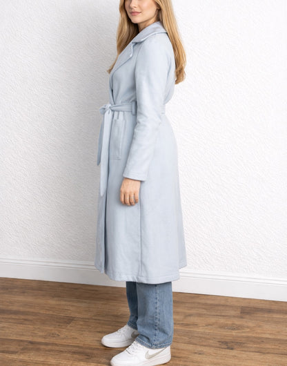 Side view of woman wearing a light blue Long Belted Coat standing against a white wall.