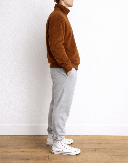 Side view of person wearing a brown fleece Bomber Jacket and gray sweatpants with white sneakers on a plain background.