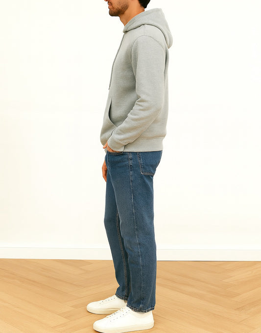 Side view of person wearing a gray hoodie and blue jeans standing against a white wall.