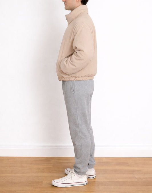 Side view of person wearing a beige Puffer Jacket and gray pants standing on a wooden floor with a white wall background.