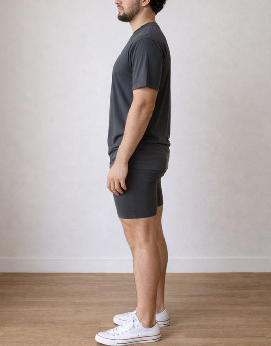 Side view of man wearing a dark gray t-shirt and biker shorts with white sneakers against a plain background.