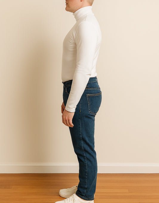 Side view of person wearing a white turtleneck and blue jeans against a beige wall.