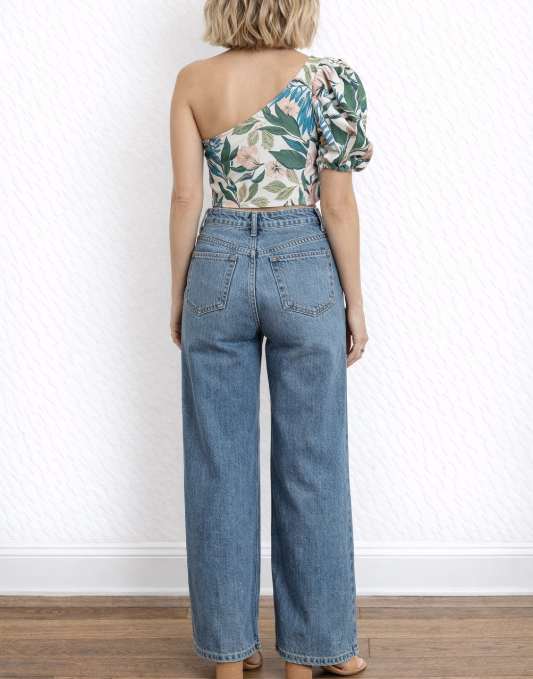 Back view of woman wearing a floral One Shoulder Puff Sleeve Crop Top and blue jeans against a white wall.