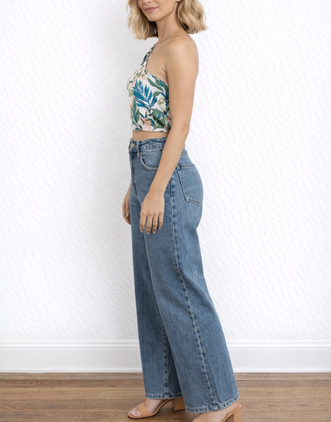 Side view of woman wearing a floral One Shoulder Puff Sleeve Crop Top and blue jeans against a white wall.