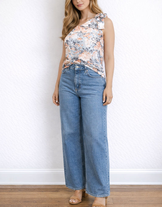 Woman wearing a floral sleeveless One Shoulder Top and blue jeans against a white wall.