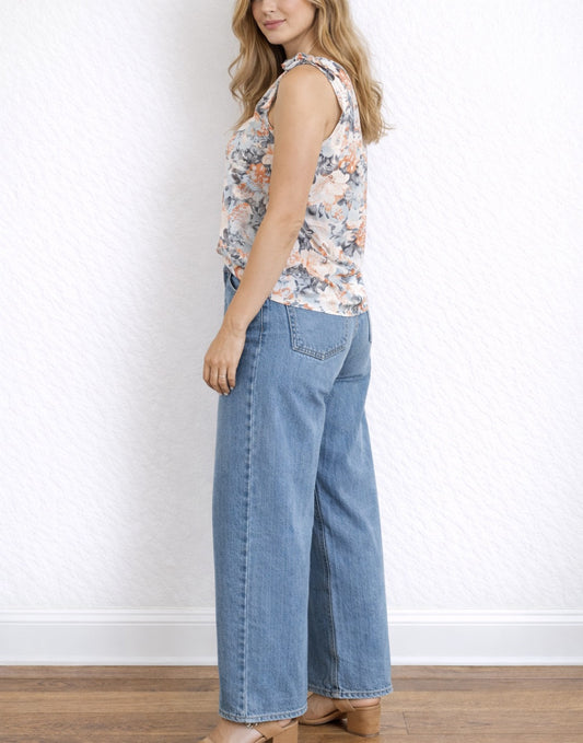 Side view of woman wearing a floral sleeveless One Shoulder Top and blue jeans against a white wall.