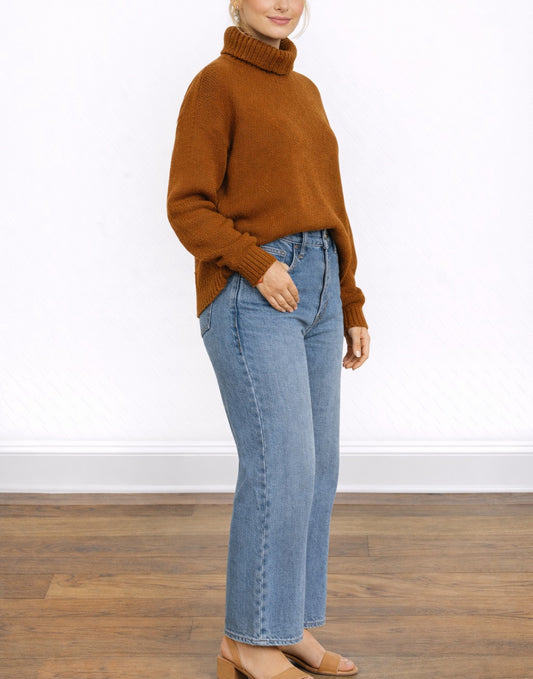Side view of woman wearing brown Oversized Turtleneck Sweater and blue jeans standing in front of a plain background.