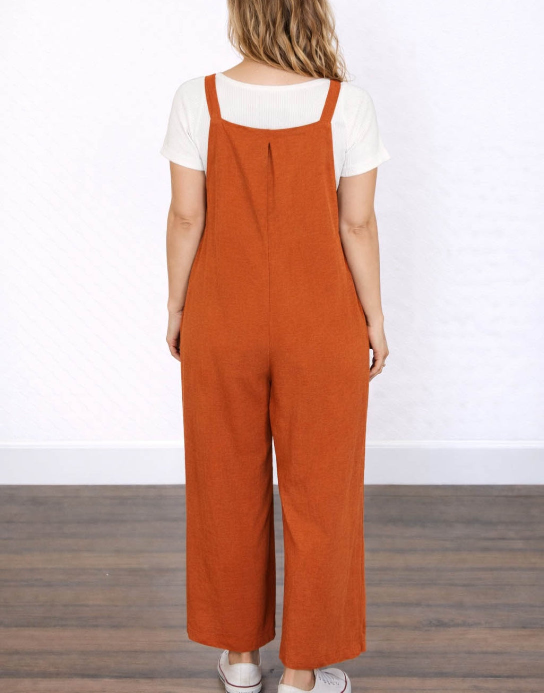 Back view of woman wearing orange Pinafore Jumpsuit with a white shirt underneath on a plain background.