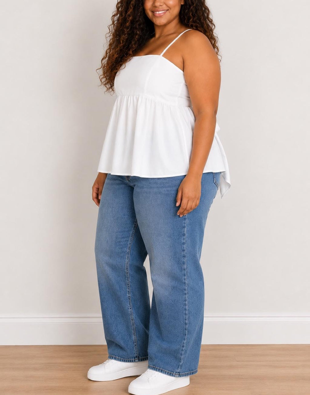 Side view of woman wearing a white Plus Size Bow Back Babydoll Cami Top and blue jeans against a plain background.