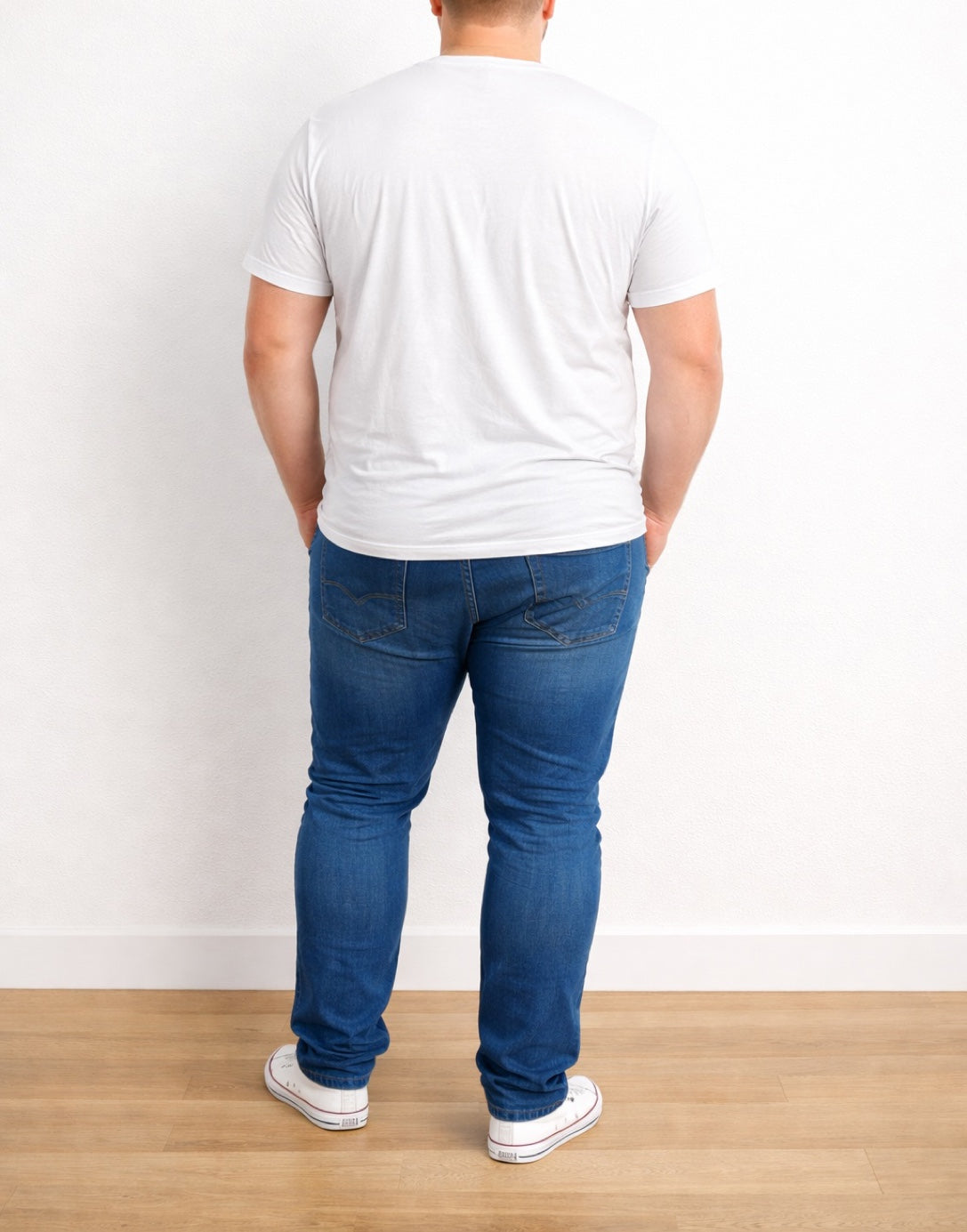 Back view of person wearing a white Plus Size Classic T-Shirt and blue jeans standing against a white wall.