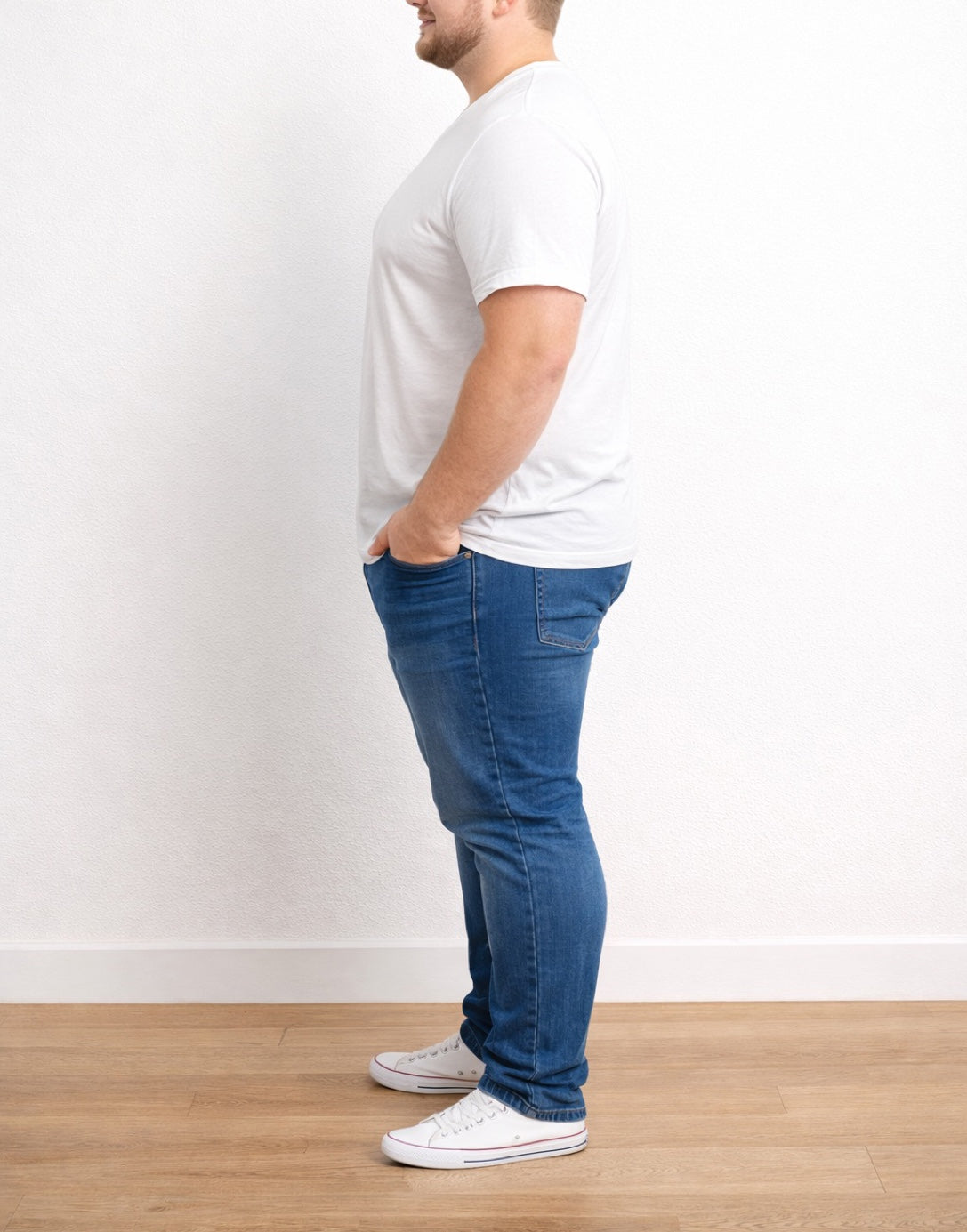 Side view of man wearing a white Plus Size Classic V-Neck Tee Shirt and blue jeans standing against a plain white wall.