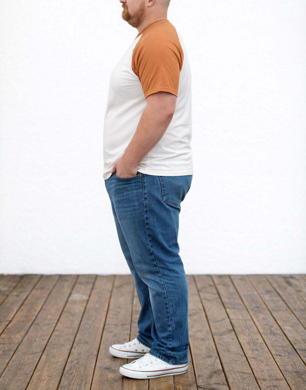 Side view of person wearing a white and orange Plus Size Crew Neck Short Sleeve Baseball Shirt, blue jeans, and white sneakers on a wooden floor.