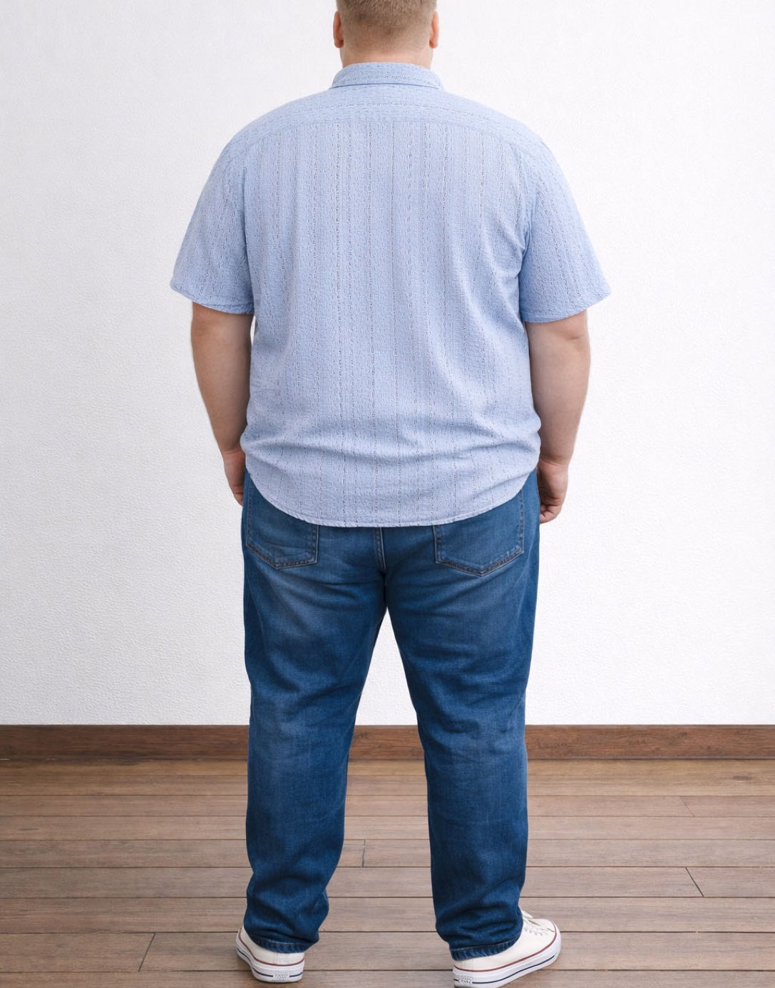 Back view of person wearing a light blue Plus Size Drop Shoulder Loose Fit Shirt and dark blue jeans standing on a wooden floor with a white wall background.
