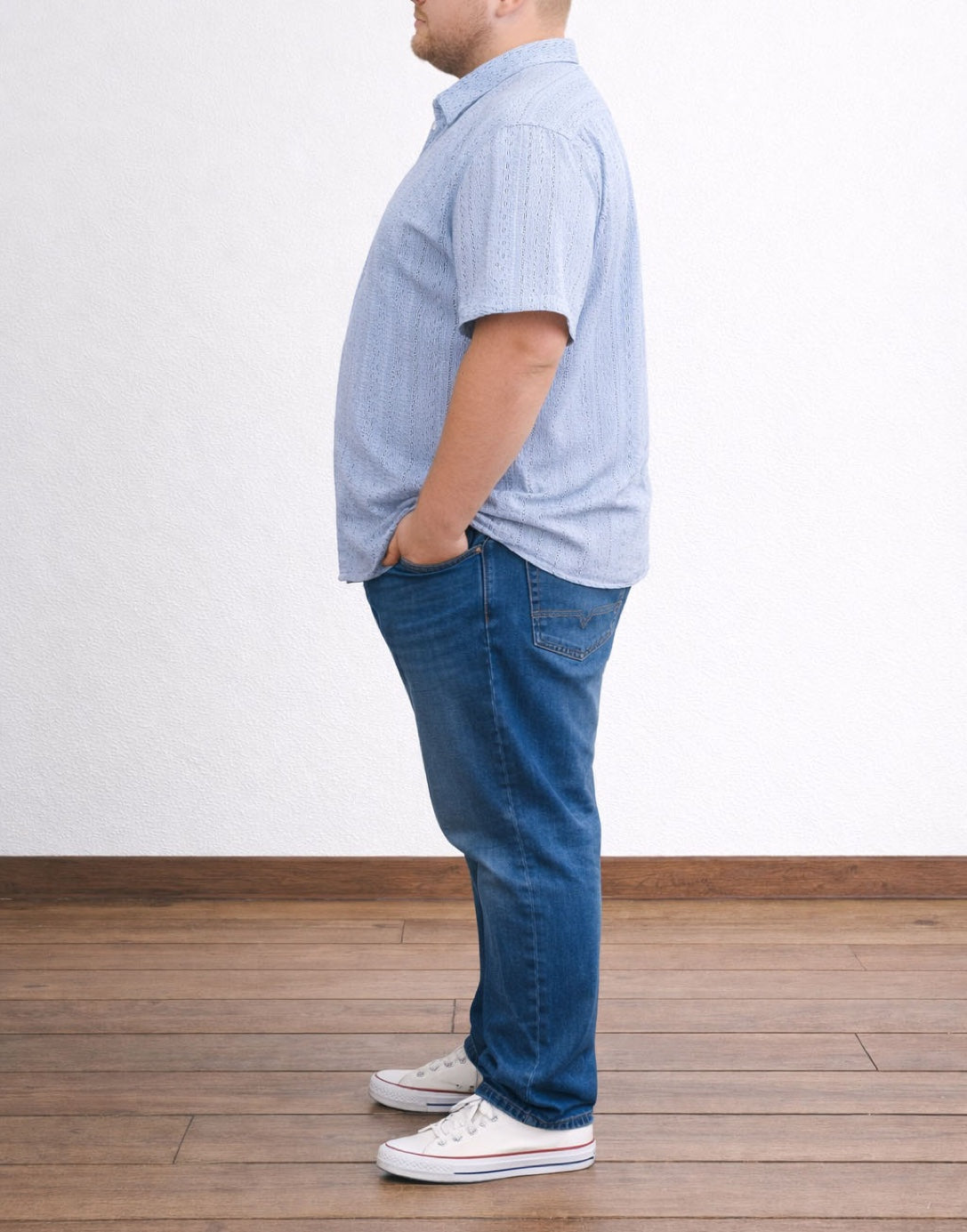 Side view of person wearing a light blue Plus Size Drop Shoulder Loose Fit Shirt and dark blue jeans standing on a wooden floor with a white wall background.