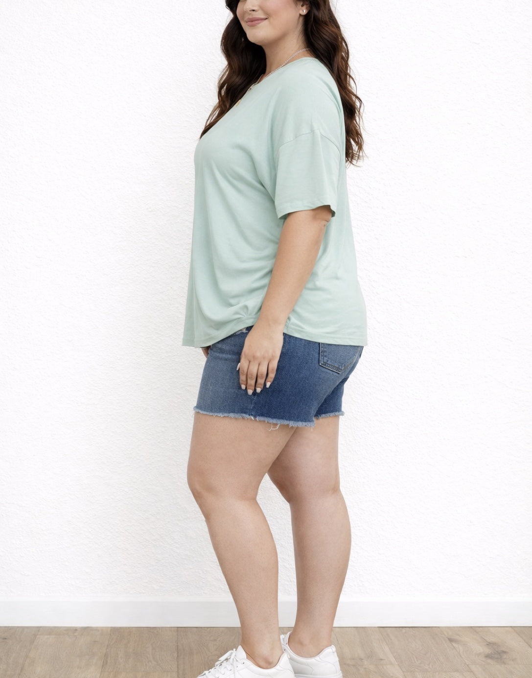 Side view of woman wearing a light green Plus Size Oversized Crew Neck Tee Shirt and denim shorts on a white background