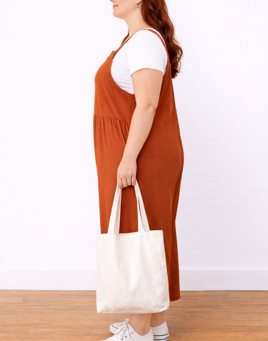 Side view of woman wearing orange Plus Size Pinafore Jumpsuit holding a white tote bag on a plain background.