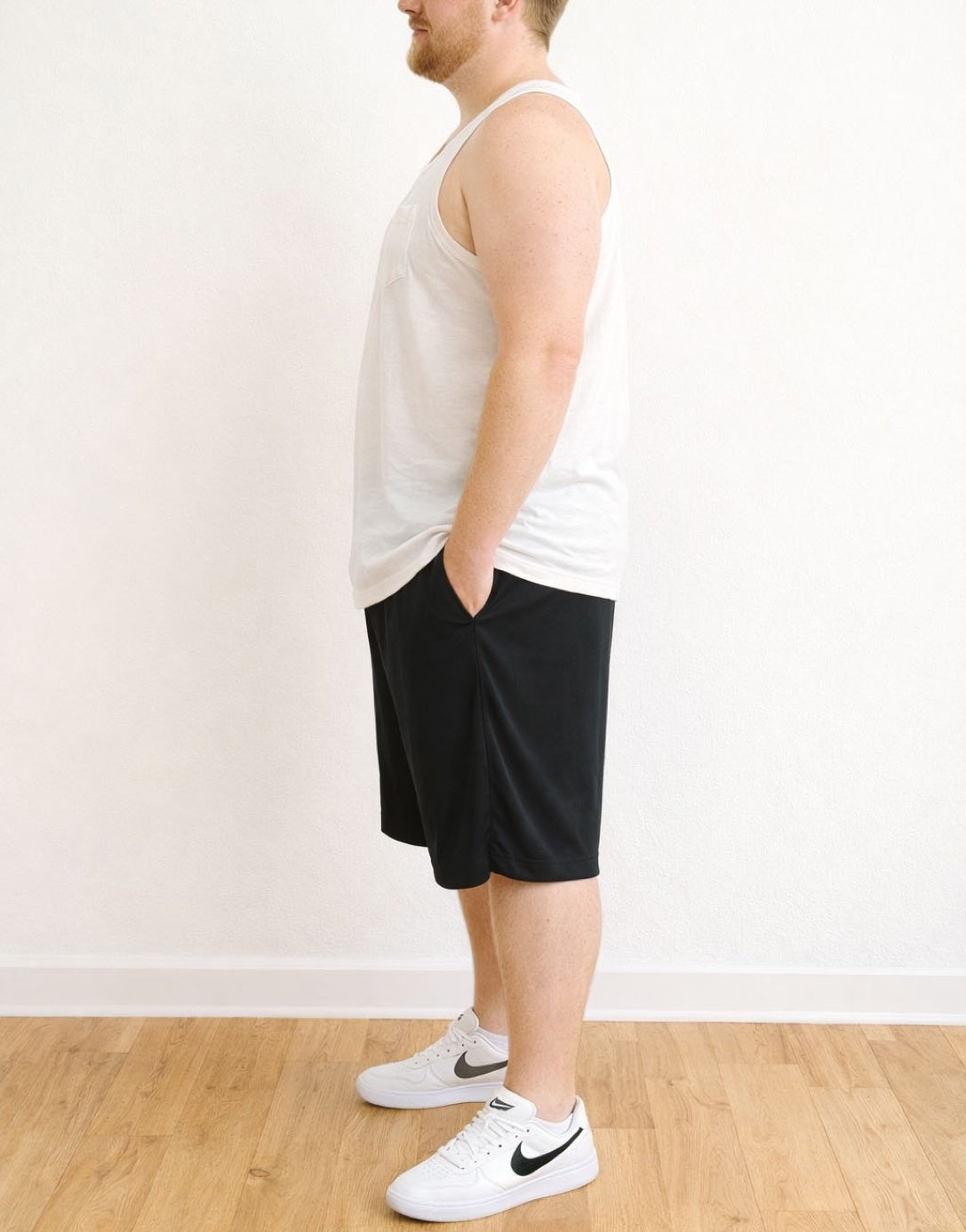 Side view of person wearing a white Plus Size Round Neck Pocket Tank Top and black shorts standing against a plain wall.