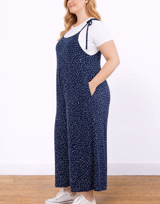 Side view of woman wearing a navy blue polka dot Plus Size Tie Shoulder Jumpsuit with a white shirt underneath on a white background..