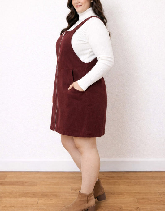 Side view of woman wearing a burgundy Plus Size Zip Front Pocket Mini Overall Dress over a white turtleneck sweater against a neutral background.