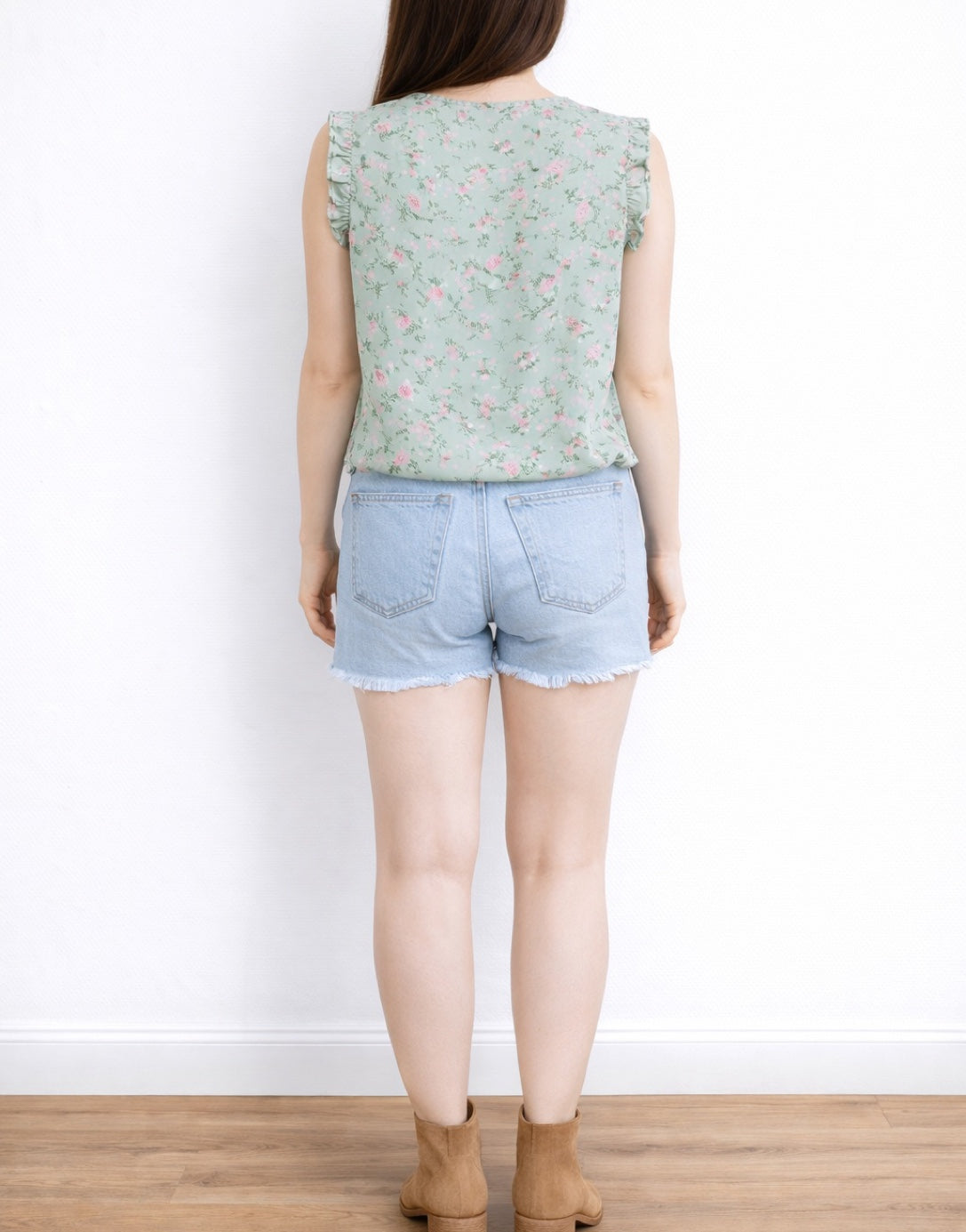 Back view of woman wearing a floral Ruffled Sleeveless Blouse and denim shorts against a white background.