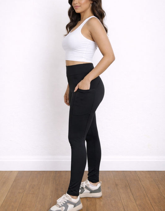 Side view of woman wearing black Side Pocket Leggings and a white crop top against a white wall.