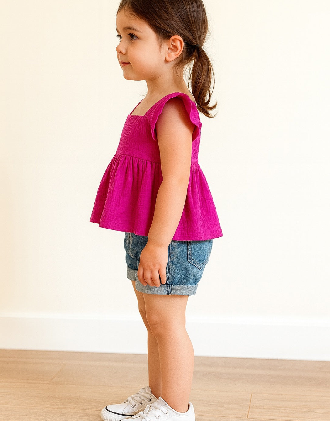 Side view of child wearing a pink Square Neck Peplum Top and denim shorts on a plain background