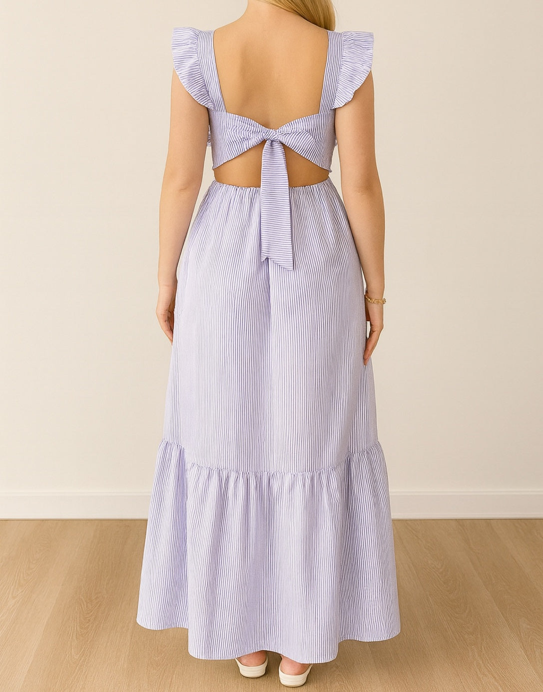 Back view of Woman wearing a light purple striped Ruffled Sleeve Maxi dress with cut-out back and tied waist on a beige background.