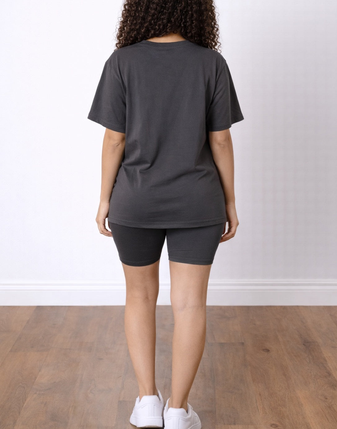 Back view of woman wearing a dark gray T-Shirt and Biker Shorts Set with white sneakers on a wooden floor.