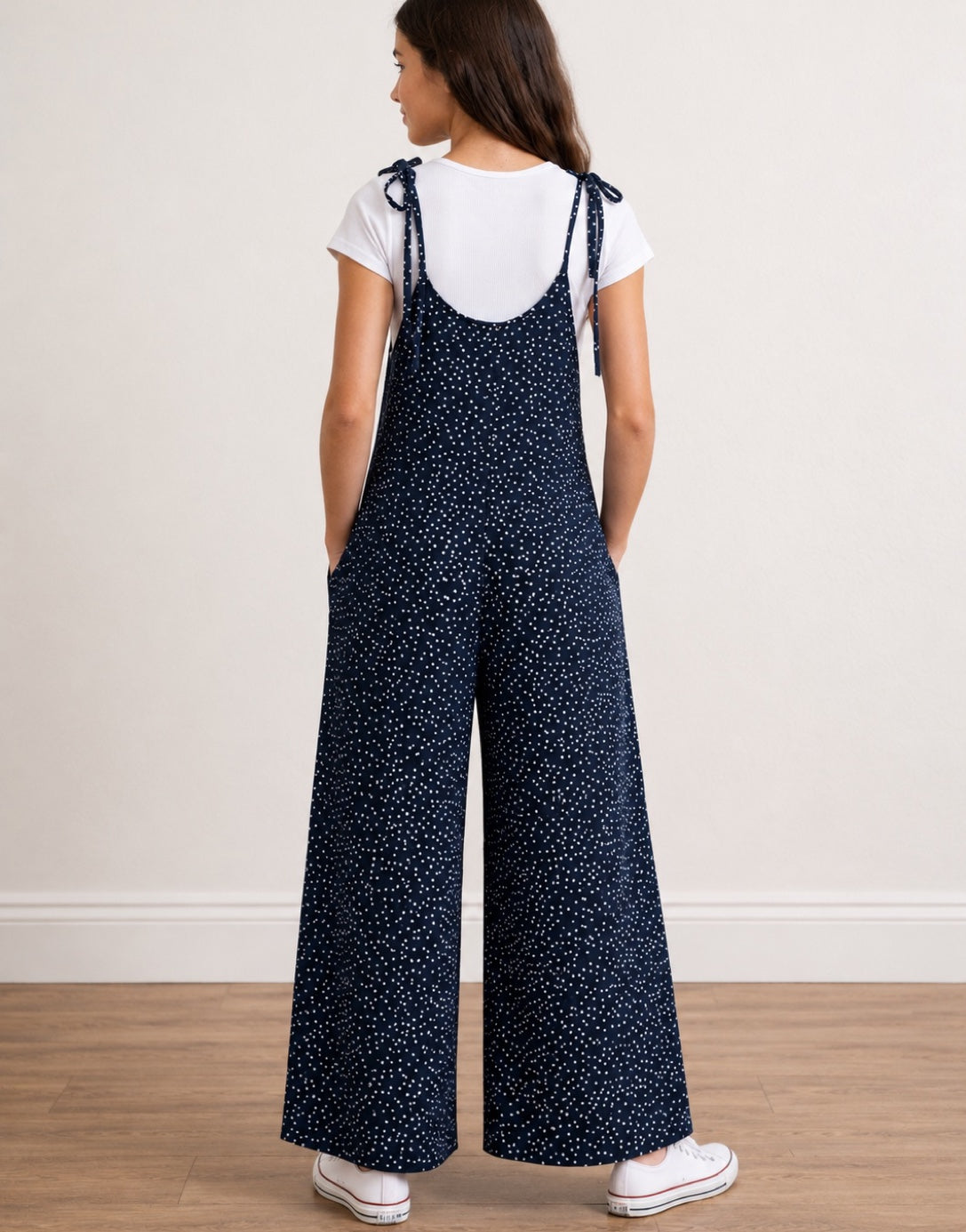 Back view of teen girl wearing navy Tie Shoulder Jumpsuit with white polka dots and white tee underneath on plain white background.