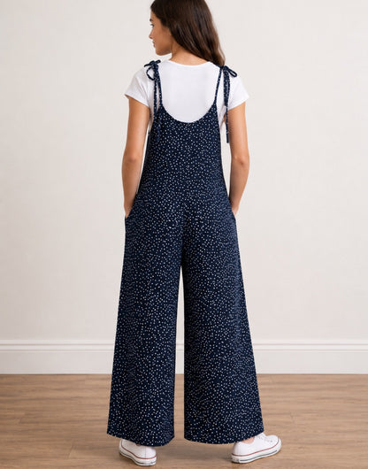 Back view of teen girl wearing navy Tie Shoulder Jumpsuit with white polka dots and white tee underneath on plain white background.