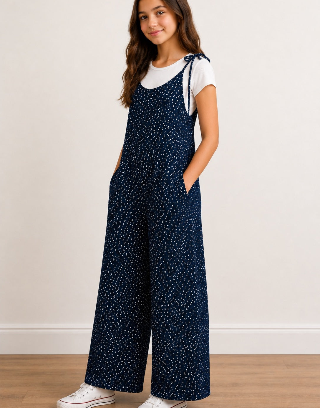 Side view of teen girl wearing navy Tie Shoulder Jumpsuit with white polka dots and white tee underneath on plain white background.