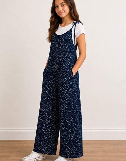Side view of teen girl wearing navy Tie Shoulder Jumpsuit with white polka dots and white tee underneath on plain white background.